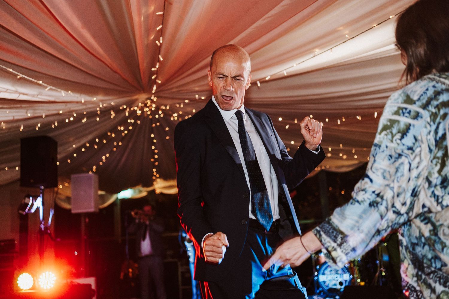Wedding guests dancing and socializing under draped fabric ceiling with string lights at an indoor reception venue.