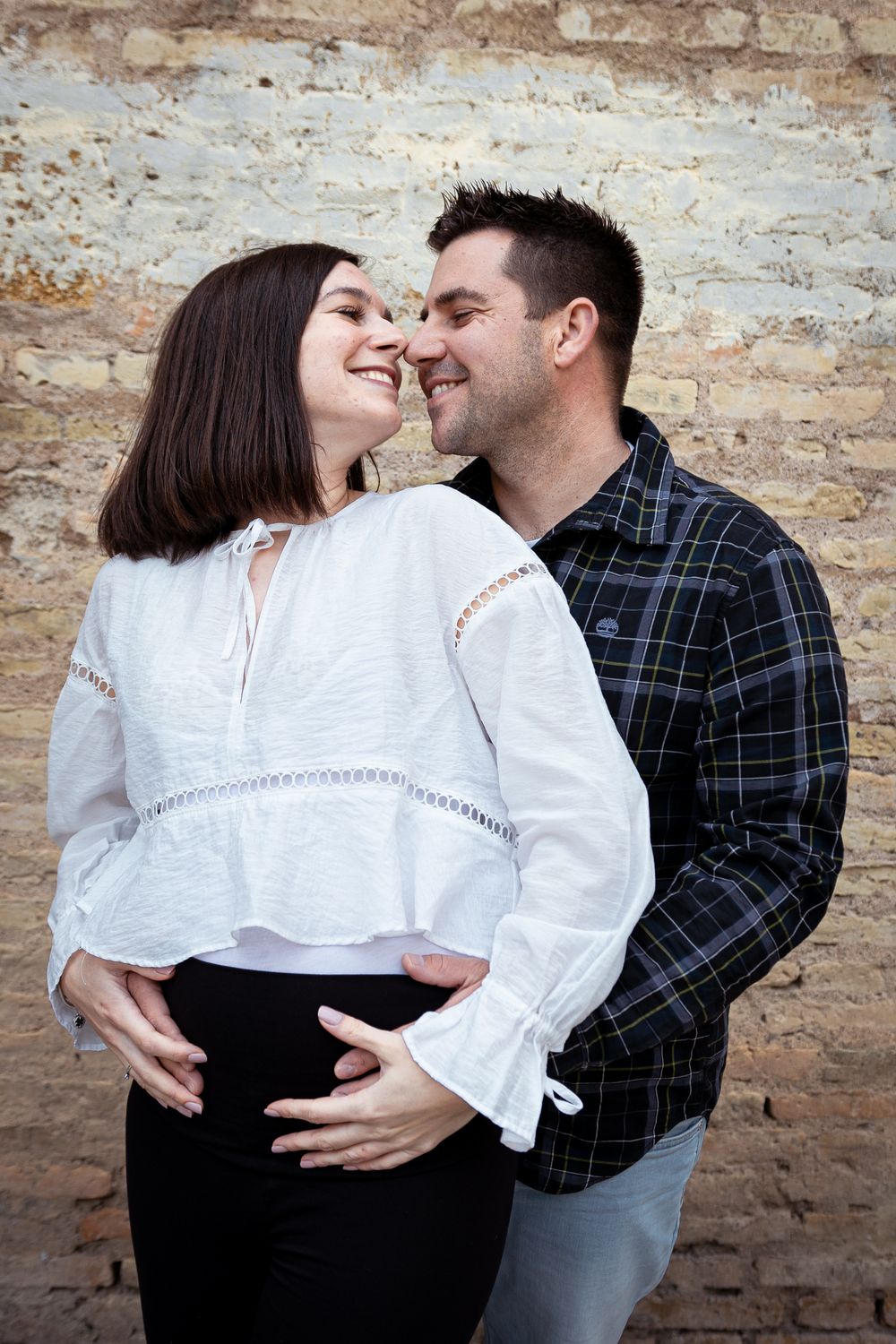 A couple in contrasting white and plaid outfits share intimate moments against a rustic brick wall.
