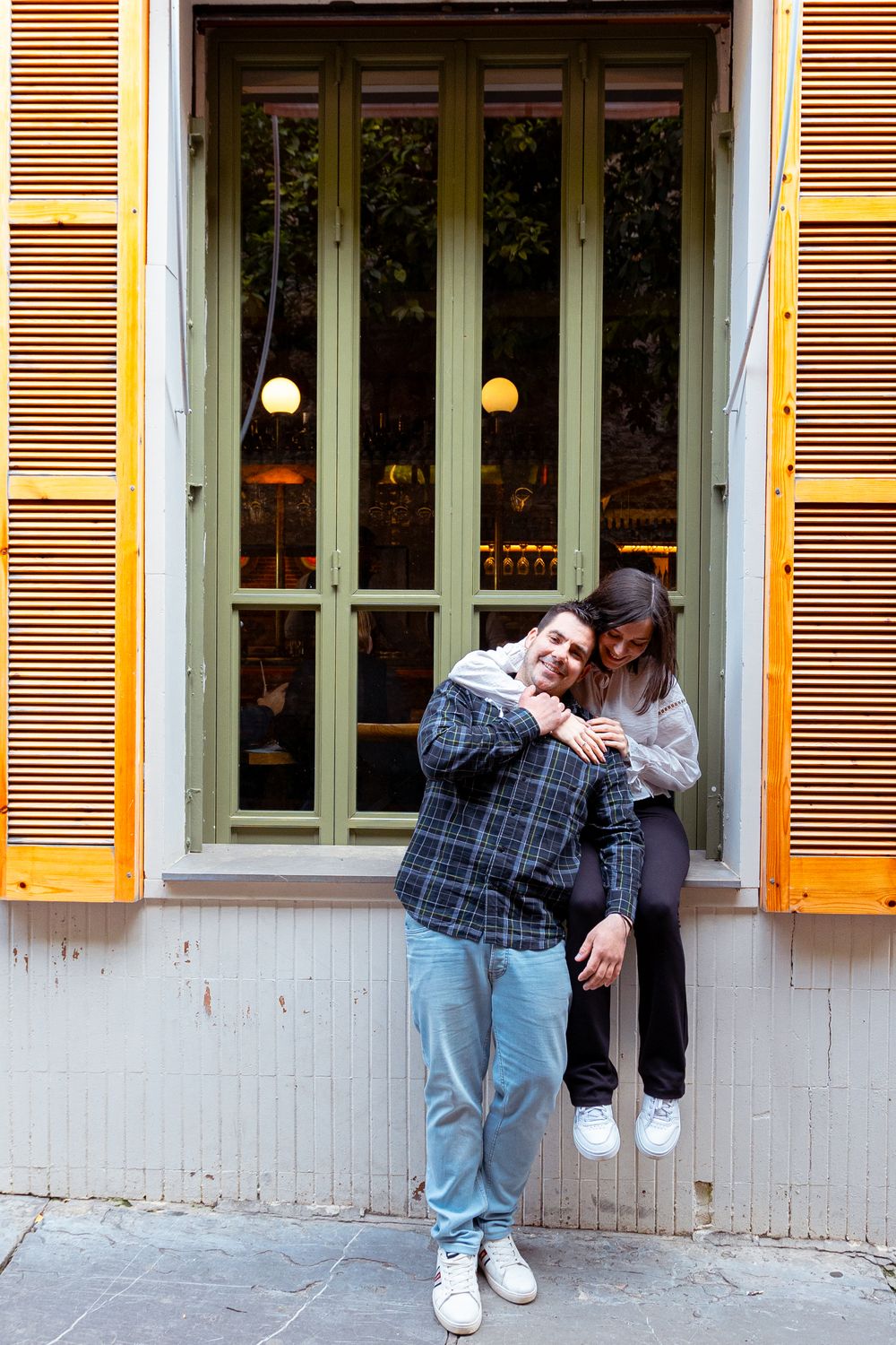 A couple in casual attire share an intimate moment against a yellow building with shuttered windows.
