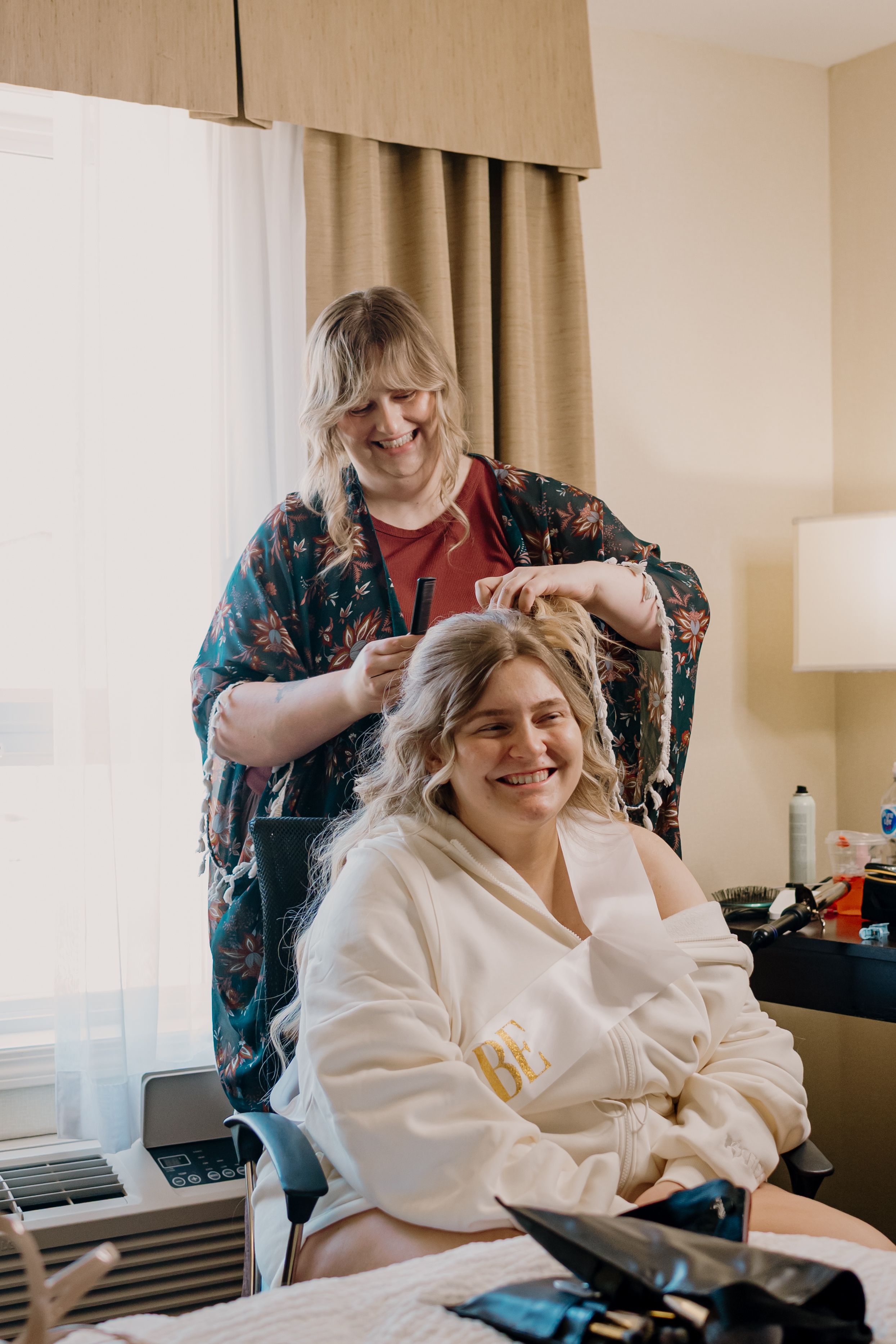A person in a white bathrobe smiles while getting their hair styled in a hotel room.