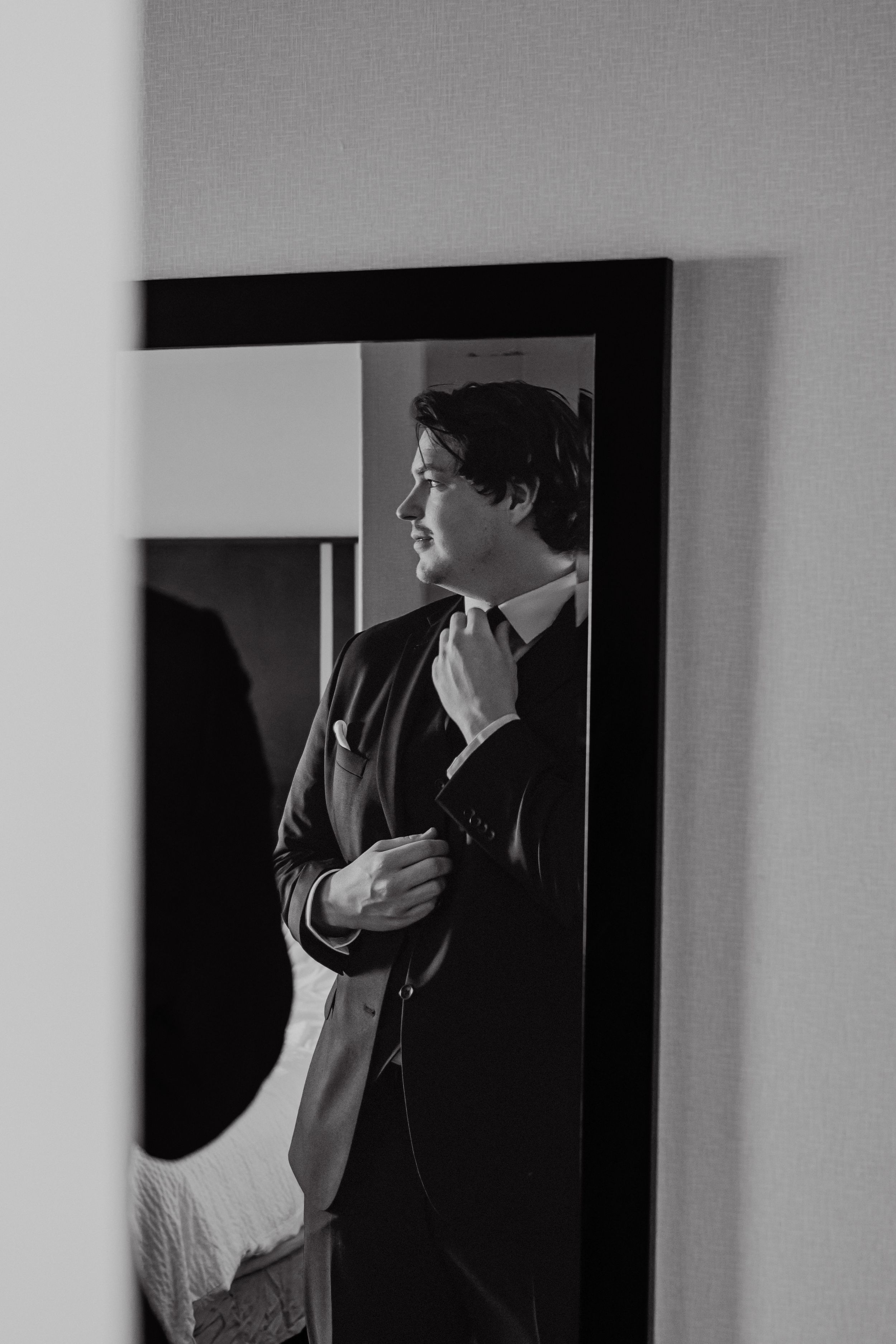 A black and white portrait of a person adjusting their tie while looking in a mirror.
