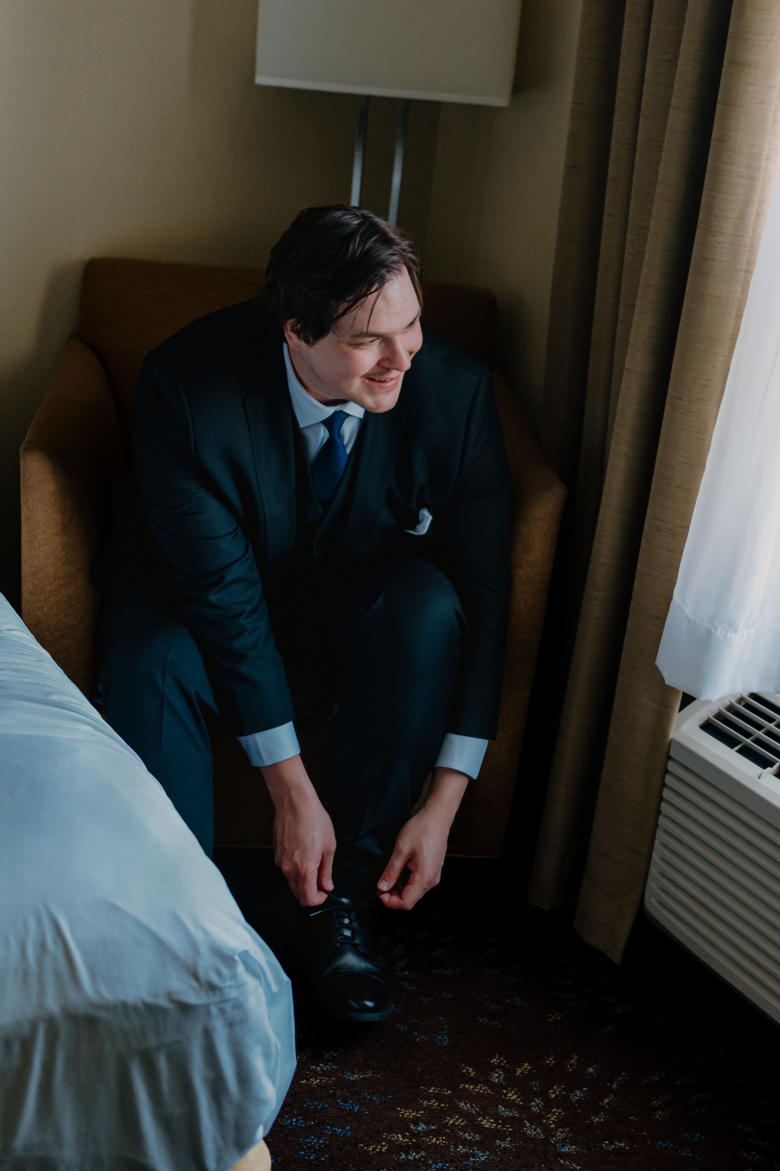 A person in a dark suit sits on a hotel room chair while putting on dress shoes.
