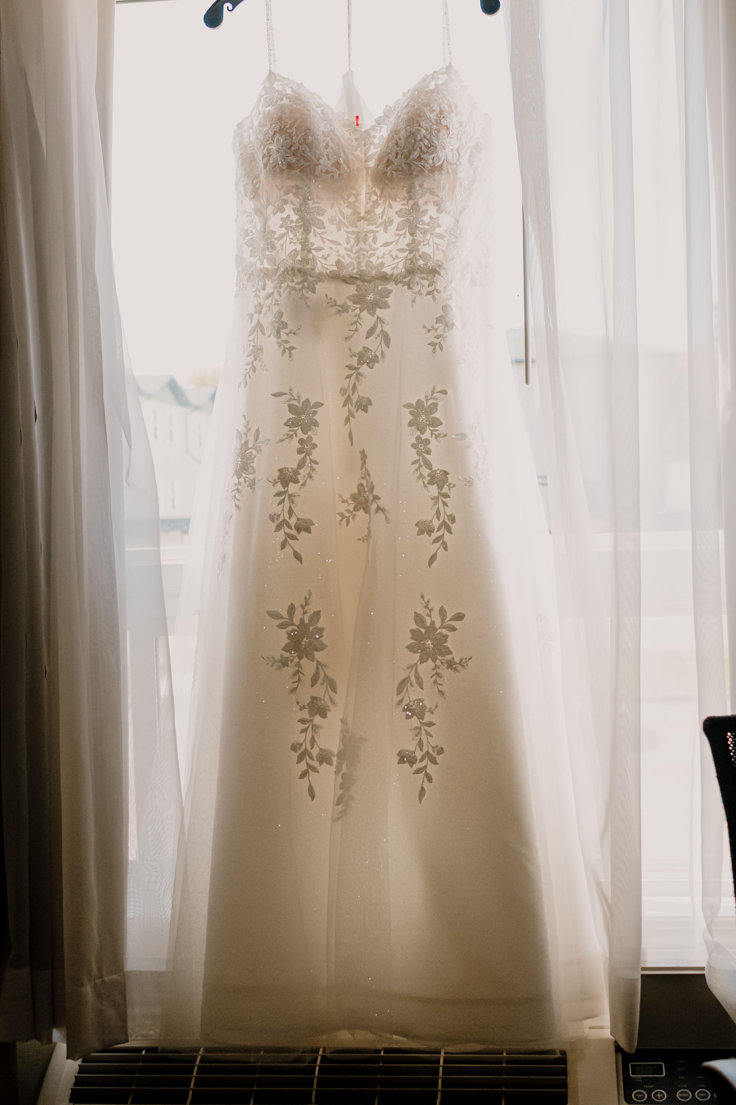 An elegant white wedding gown with floral embroidery hangs against a window with sheer curtains.
