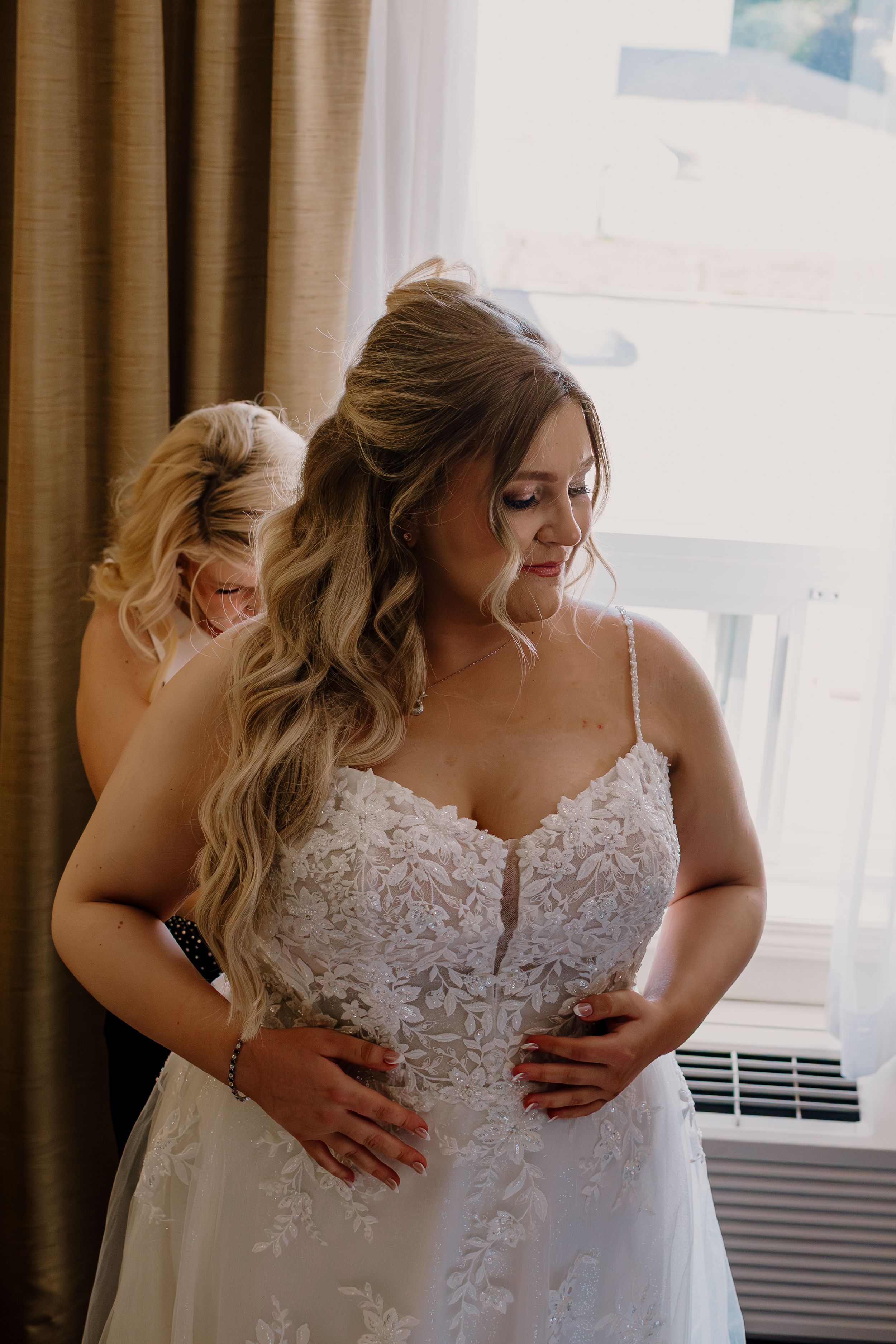 A bride in a lace wedding dress with long wavy hair gets ready in a sunlit room.