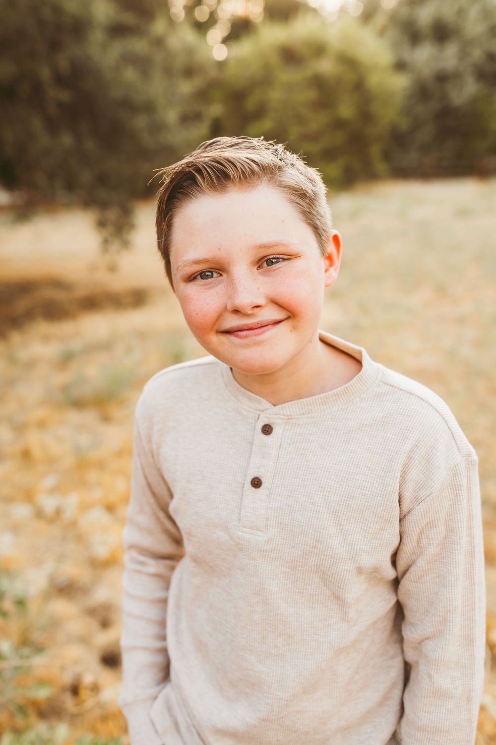 A child in a gray sweater smiles warmly while sitting outdoors during a golden hour photoshoot.