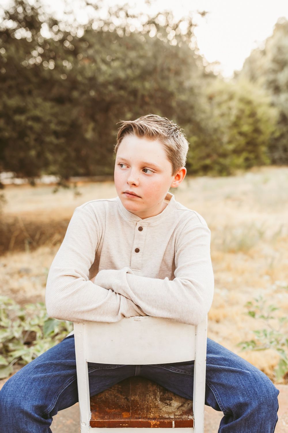 Young person in beige henley shirt and jeans sits casually on white chair in outdoor setting during golden hour.