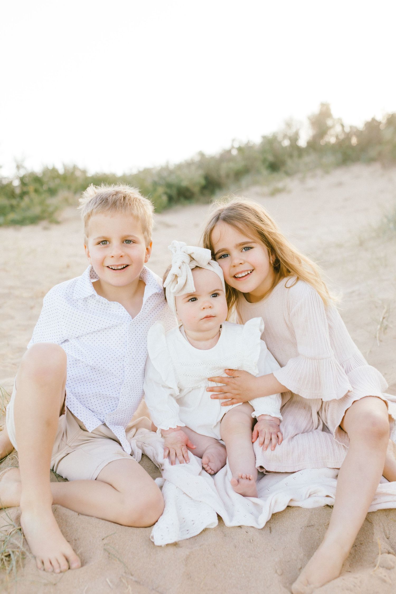 Children in white outfits sit together on sandy beach during sunset photoshoot.