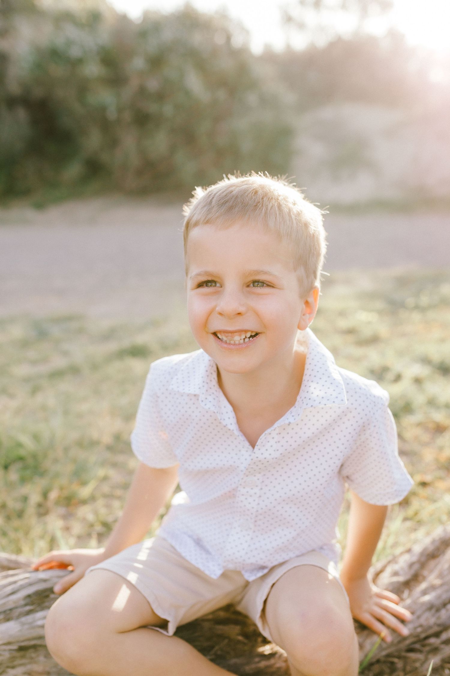 A child wearing a white polka dot shirt sits on a log during golden hour with sunlight creating a warm glow.