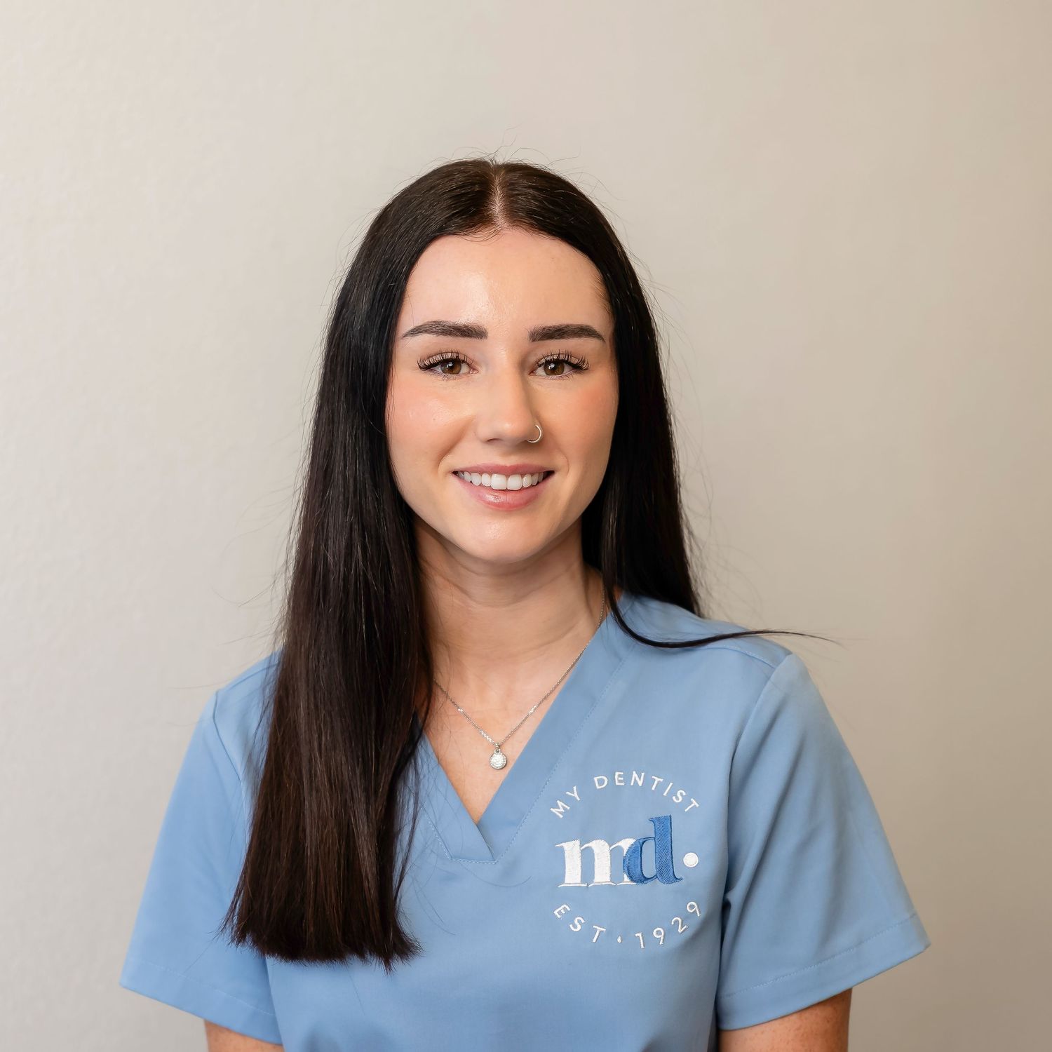 Healthcare professional in light blue scrubs smiles while posing against neutral background.