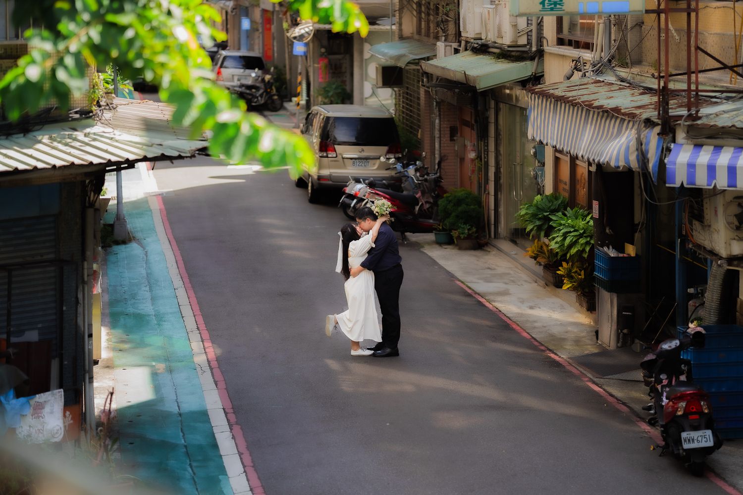 A couple in wedding attire embraces in the middle of a quiet urban alley with green foliage and buildings on both sides.