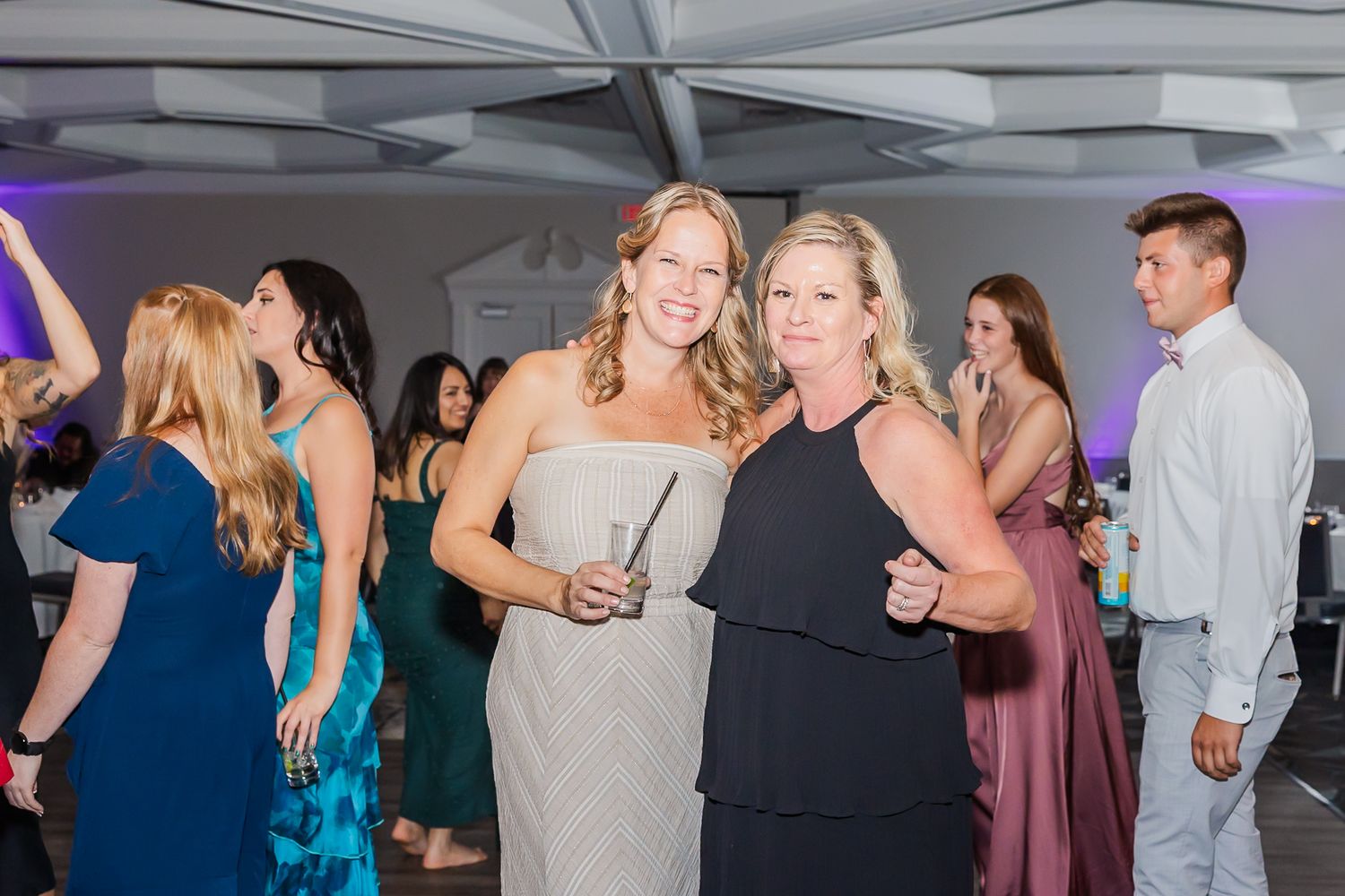 Wedding guests dance and celebrate on the dance floor at an indoor reception with purple lighting.
