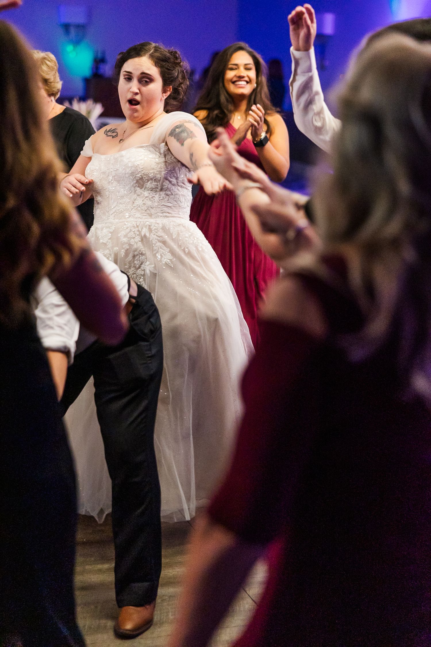 A bride in a white dress dances energetically with guests on a dimly lit dance floor at a wedding reception.