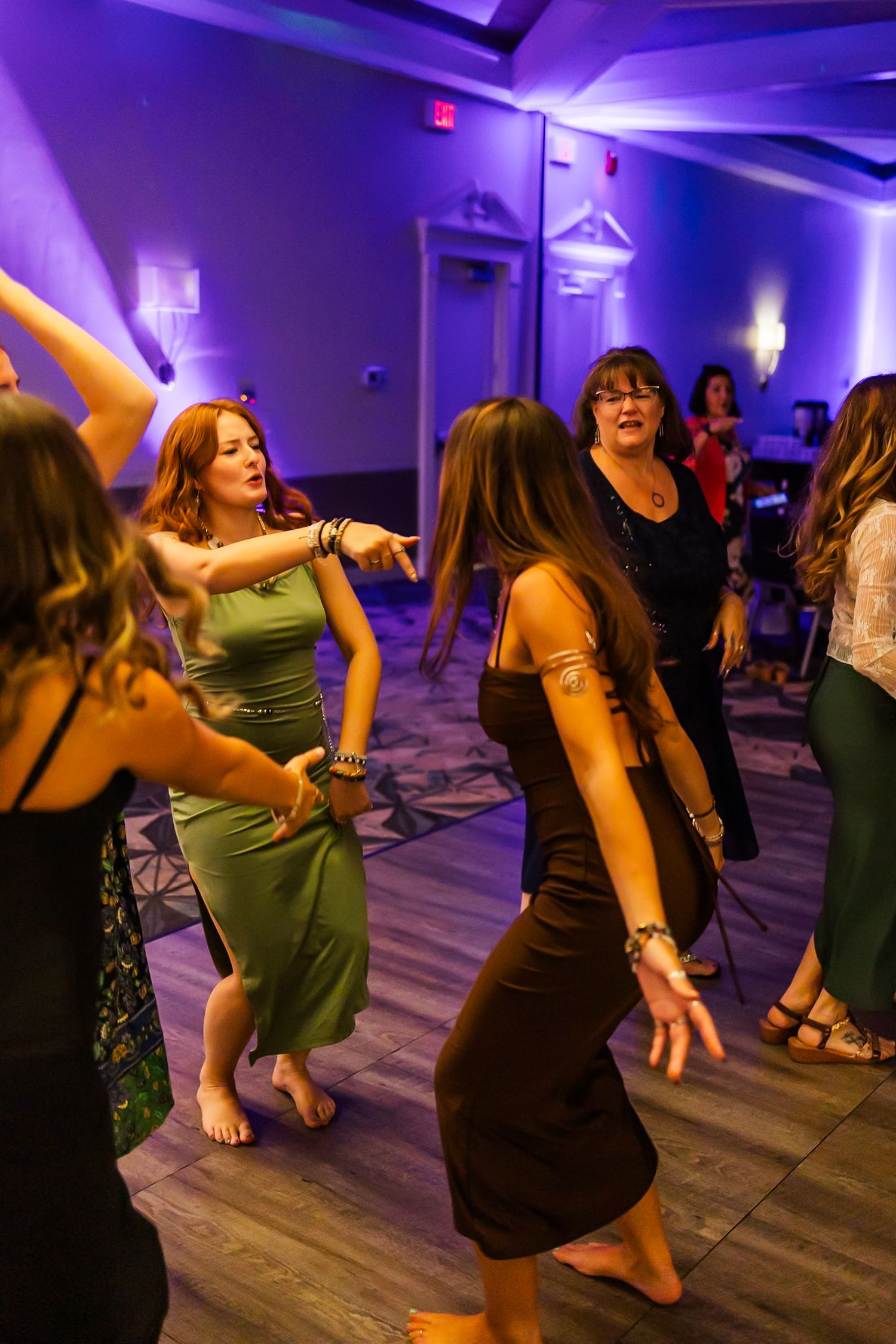 Group dance class participants move energetically in a purple-lit studio with hardwood floors during an evening session.