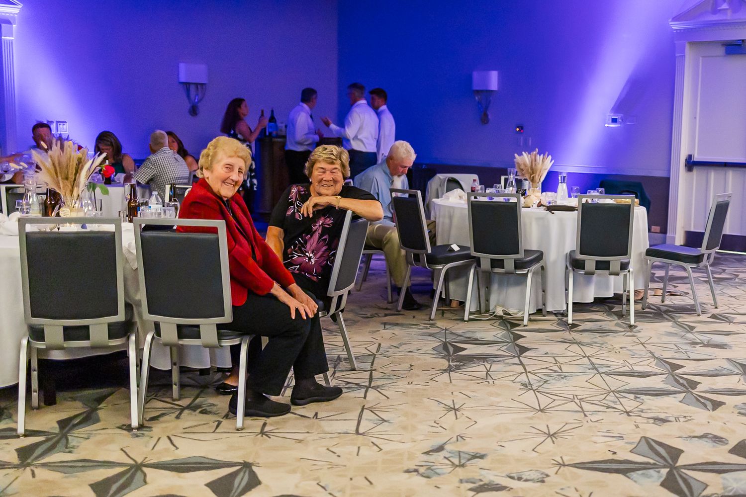 Empty chairs arranged in rows face a stage area in a venue with purple ambient lighting and patterned flooring.