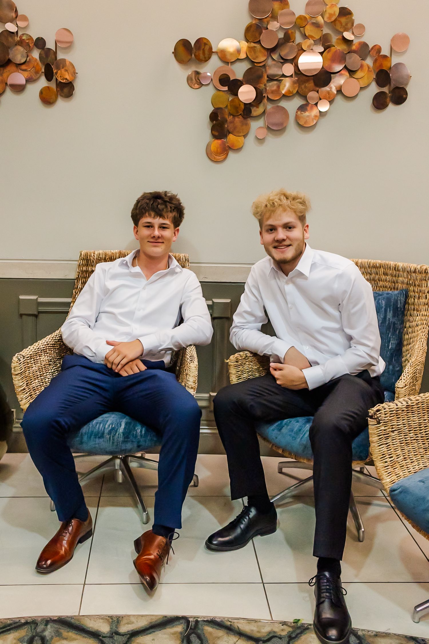 Two young men in formal attire sitting on wicker chairs against a wall decorated with copper circles.