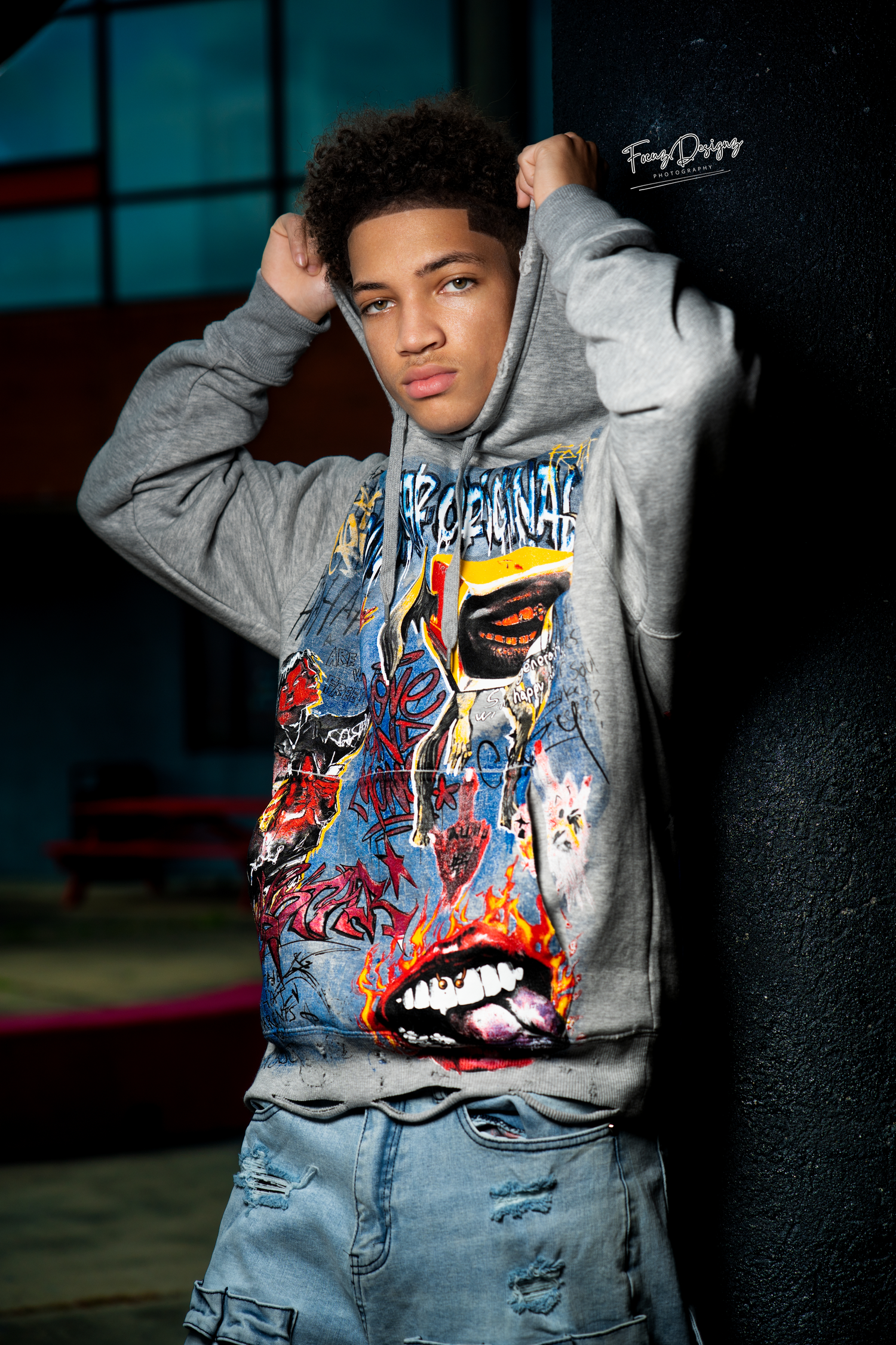 Model wearing a gray sweatshirt with colorful graffiti-style artwork and blue jeans in a dark moody setting.