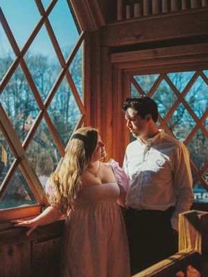 A romantic couple stands by a window with diamond-paned glass in a warm, vintage-style indoor setting at sunset.