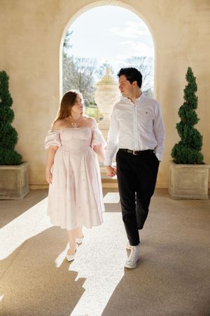A couple in coordinated outfits takes a romantic stroll through an arched Mediterranean-style corridor.