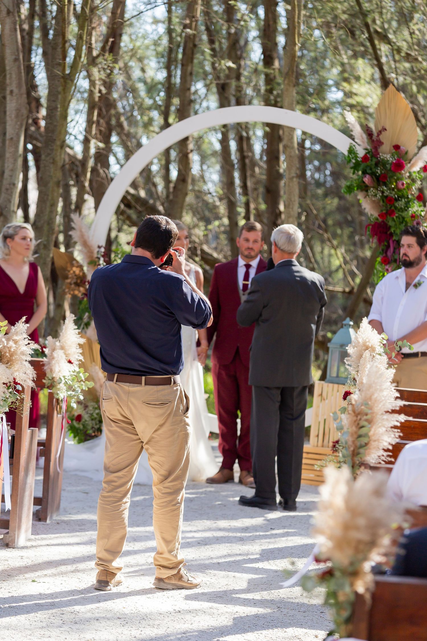 Wedding ceremony outdoors in a wooded area with white arch and pampas grass decorations during sunny day.