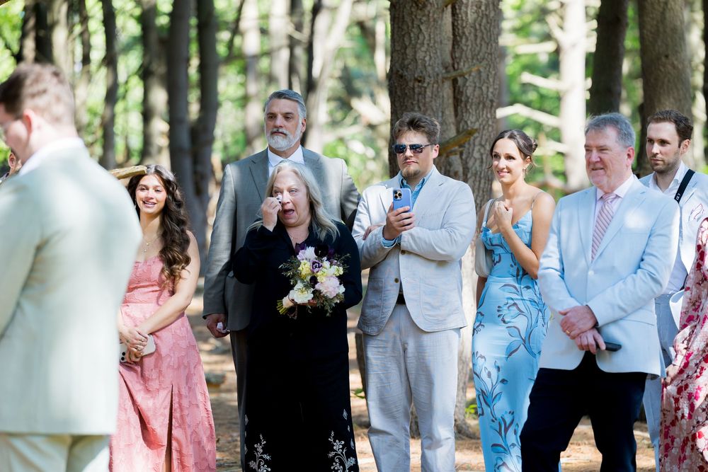 Wedding guests mingle outdoors at a garden reception beneath tall trees on a sunny afternoon.