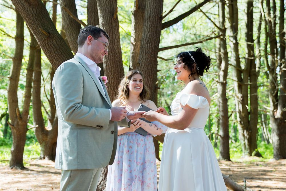 A wedding ceremony takes place in a sunlit forest with an officiant and couple exchanging rings.