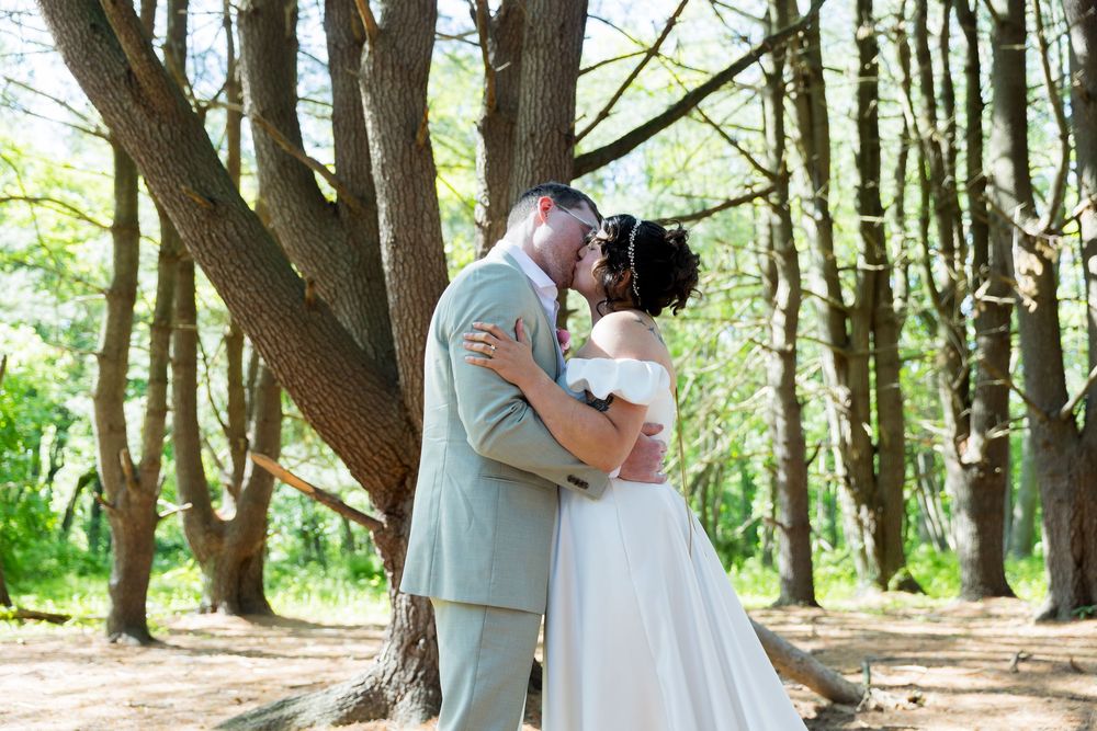 A romantic woodland scene shows a couple sharing a kiss under tall trees on their wedding day.