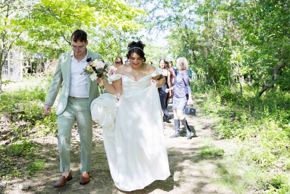 A couple walks down a wooded path during an outdoor wedding ceremony, with the bride wearing a flowing white dress.