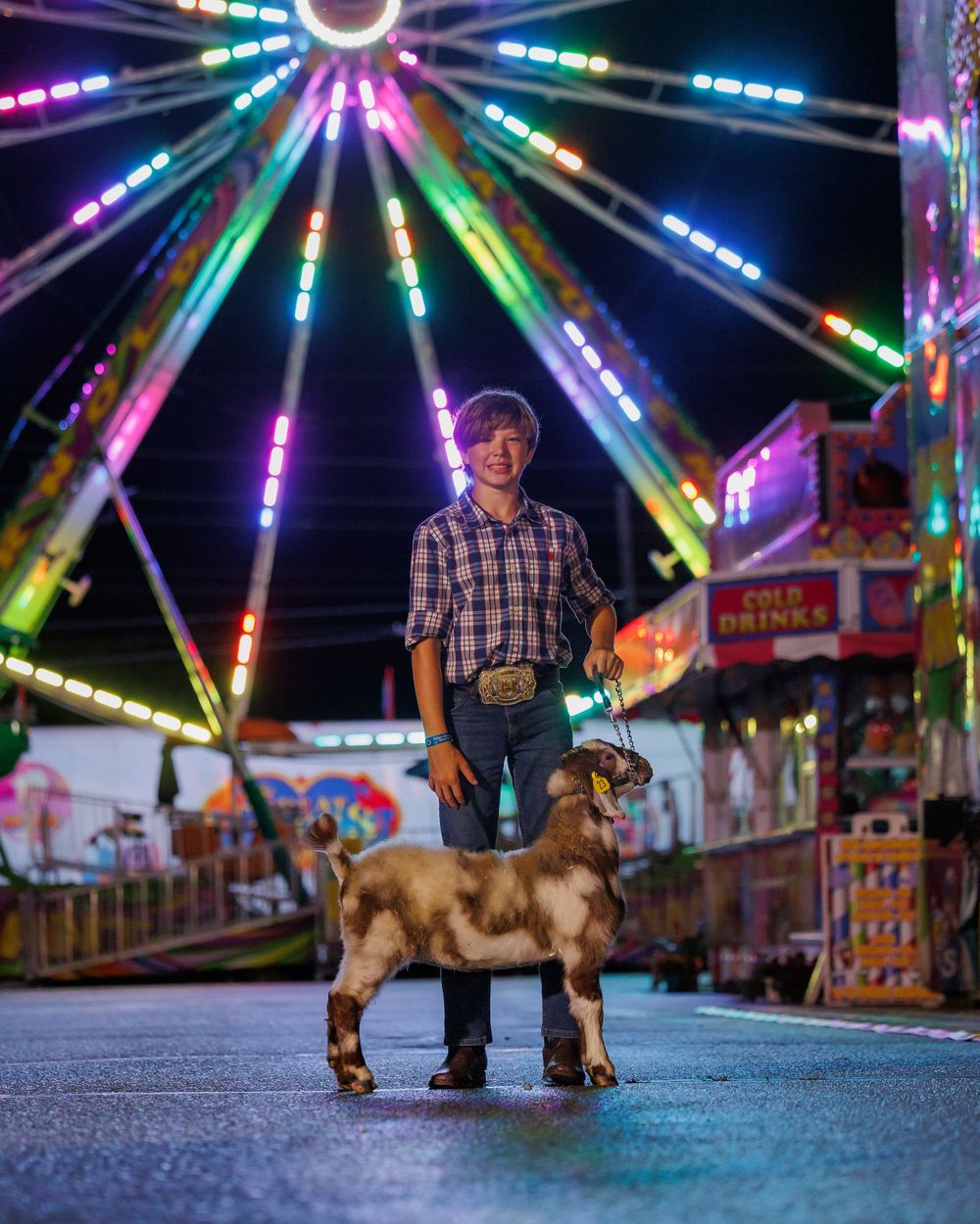 A person stands with their brown dog beneath a colorful illuminated ferris wheel at a carnival at night.