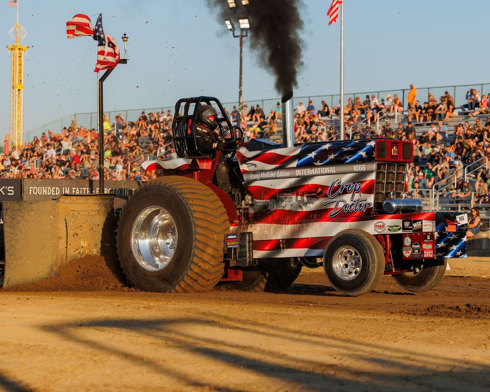 A tractor pull competition featuring an American flag-themed pulling vehicle spewing black smoke on a dirt track.