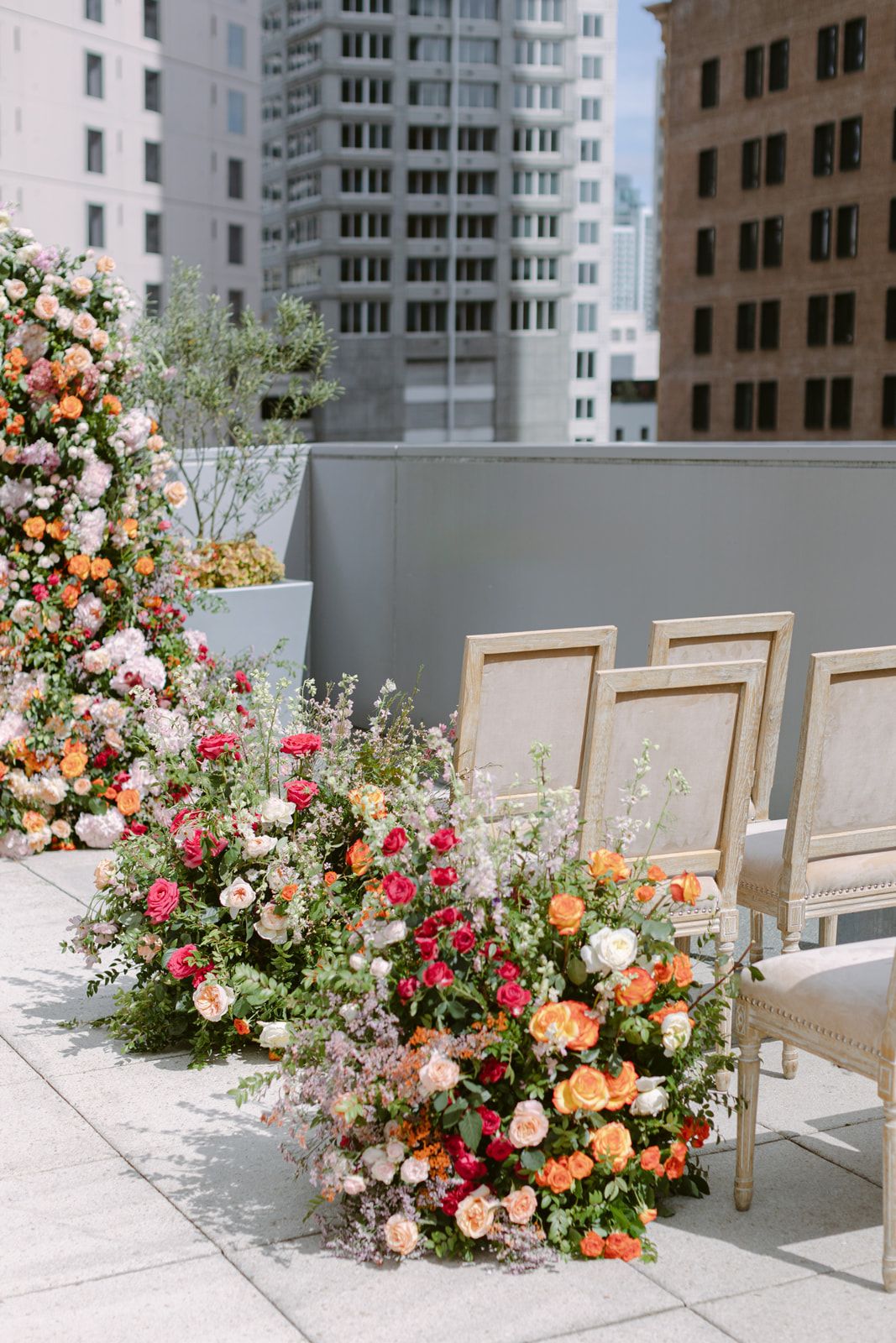 Elegant rooftop wedding ceremony setup with vintage chairs and lush floral arrangements in peach and coral against city buildings.