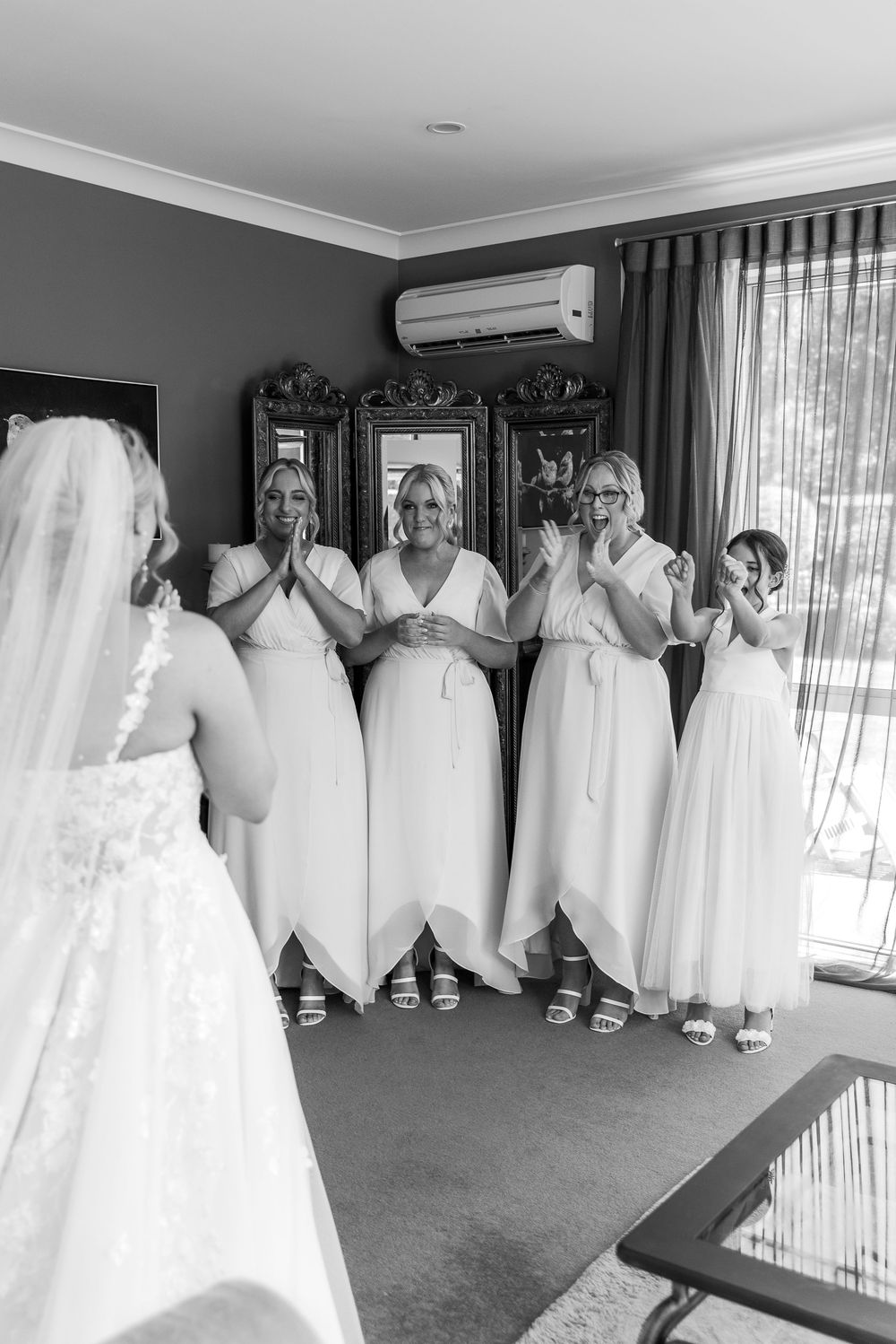 Bridesmaids in white dresses react with joy while viewing the bride in a formal getting-ready room.