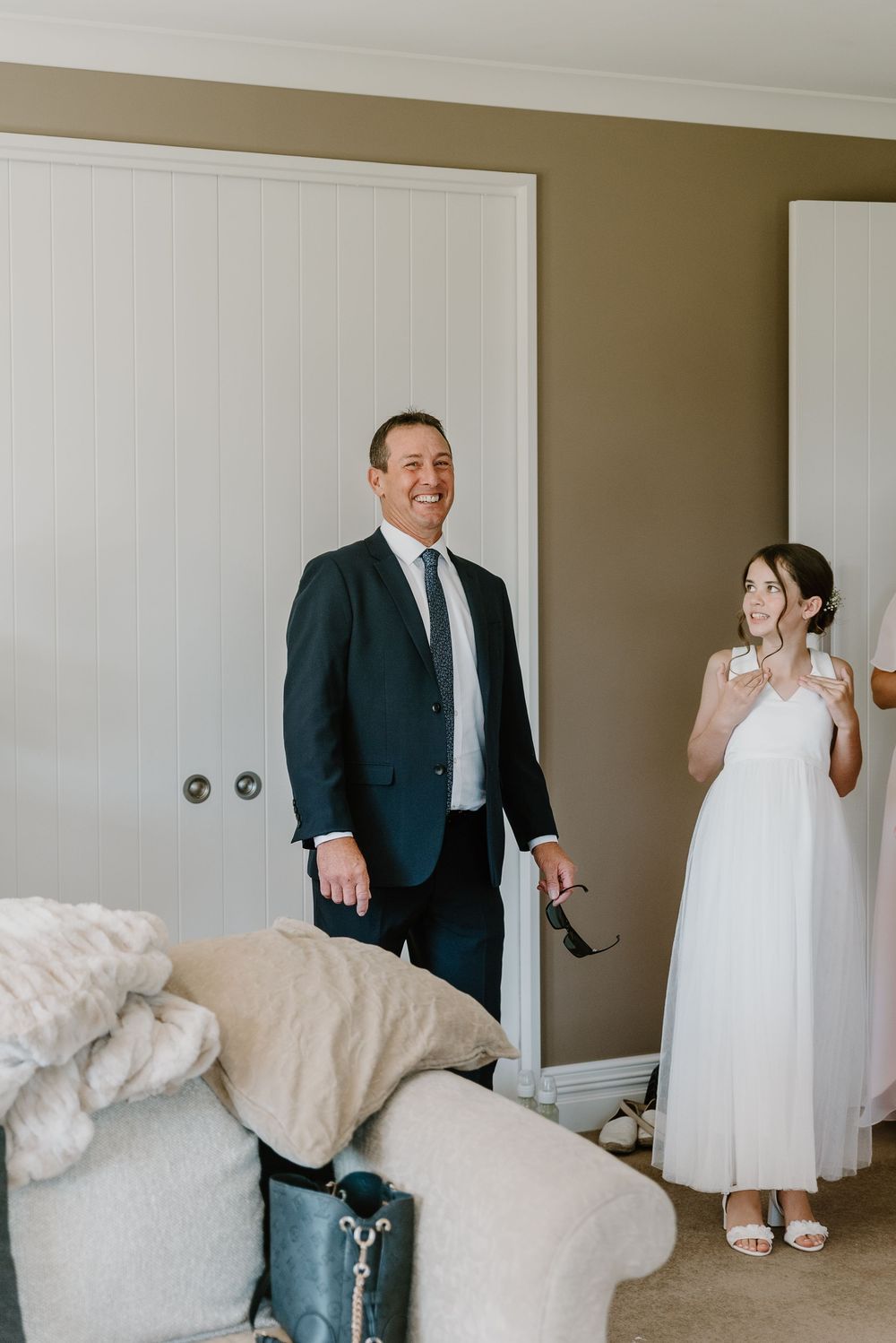 Bride and father share emotional first look moment in a sunlit room with white furnishings.