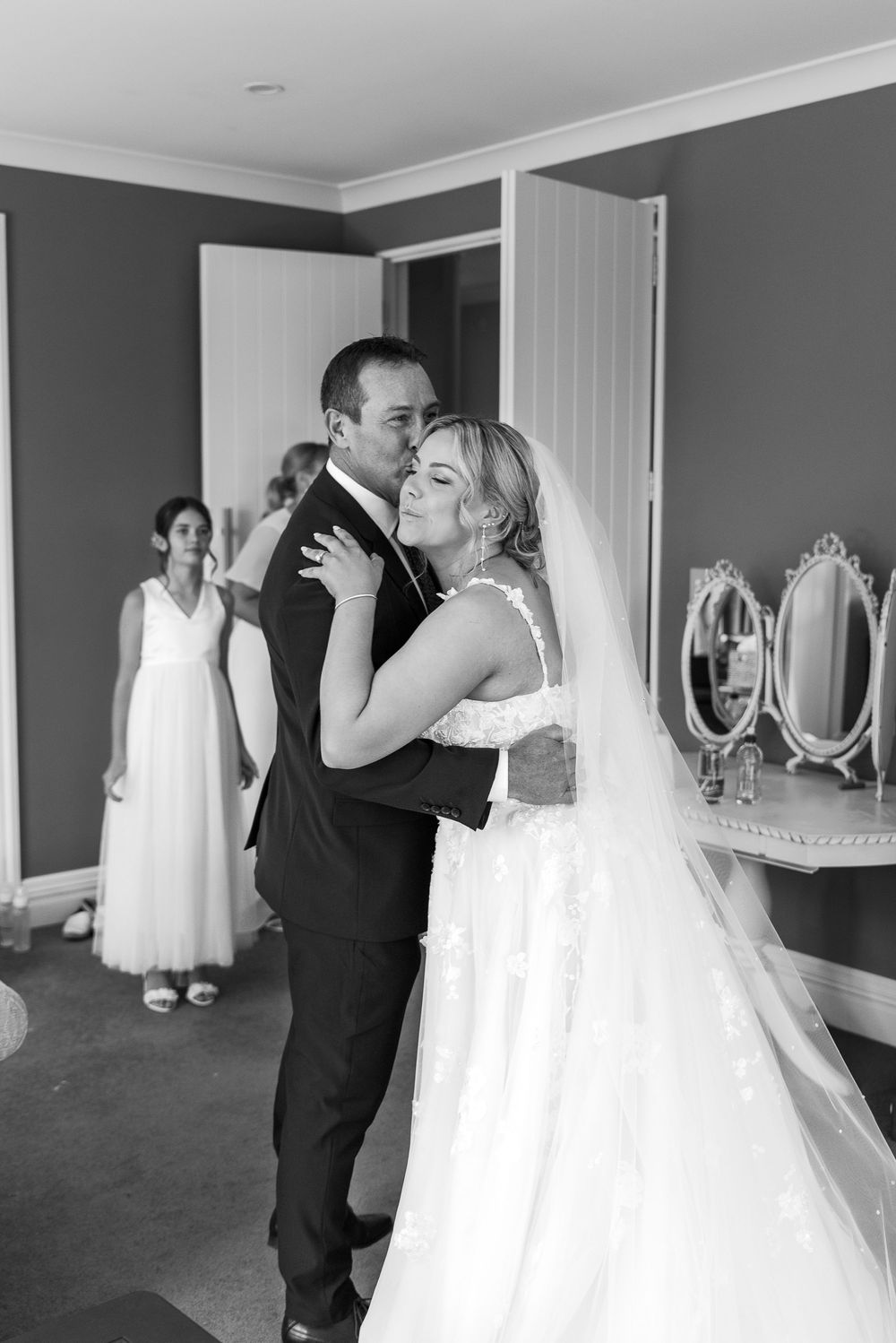 Wedding preparation scene in a gray-walled room with white bridal gown visible in mirror reflections.