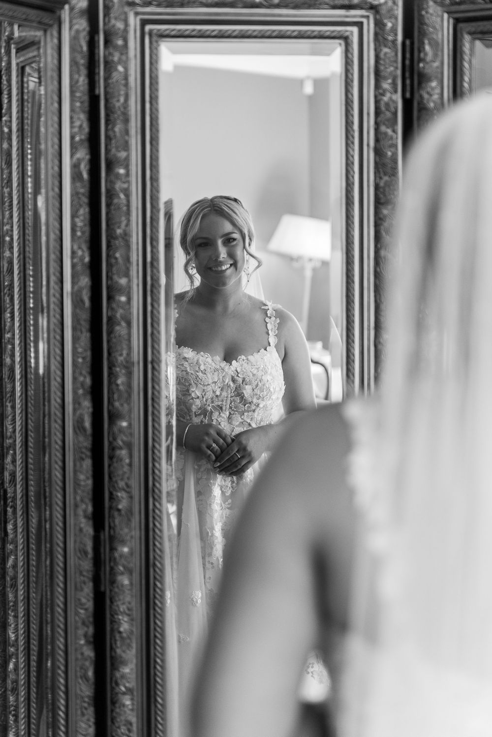 A black and white reflection of a bride in an ornate mirror wearing a lace wedding gown.