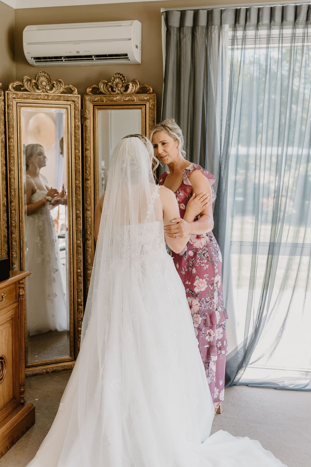 A wedding dress hangs gracefully in a room with ornate gold mirrors and sheer curtains.