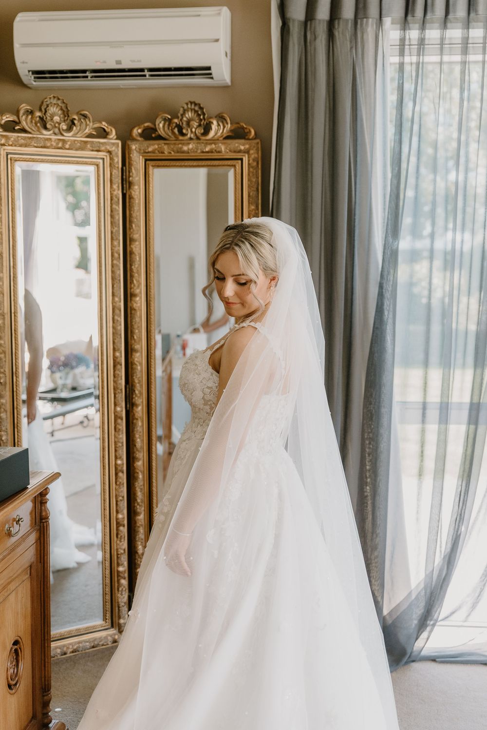 A bride in a white wedding gown poses by ornate gold mirrors in a sunlit room.