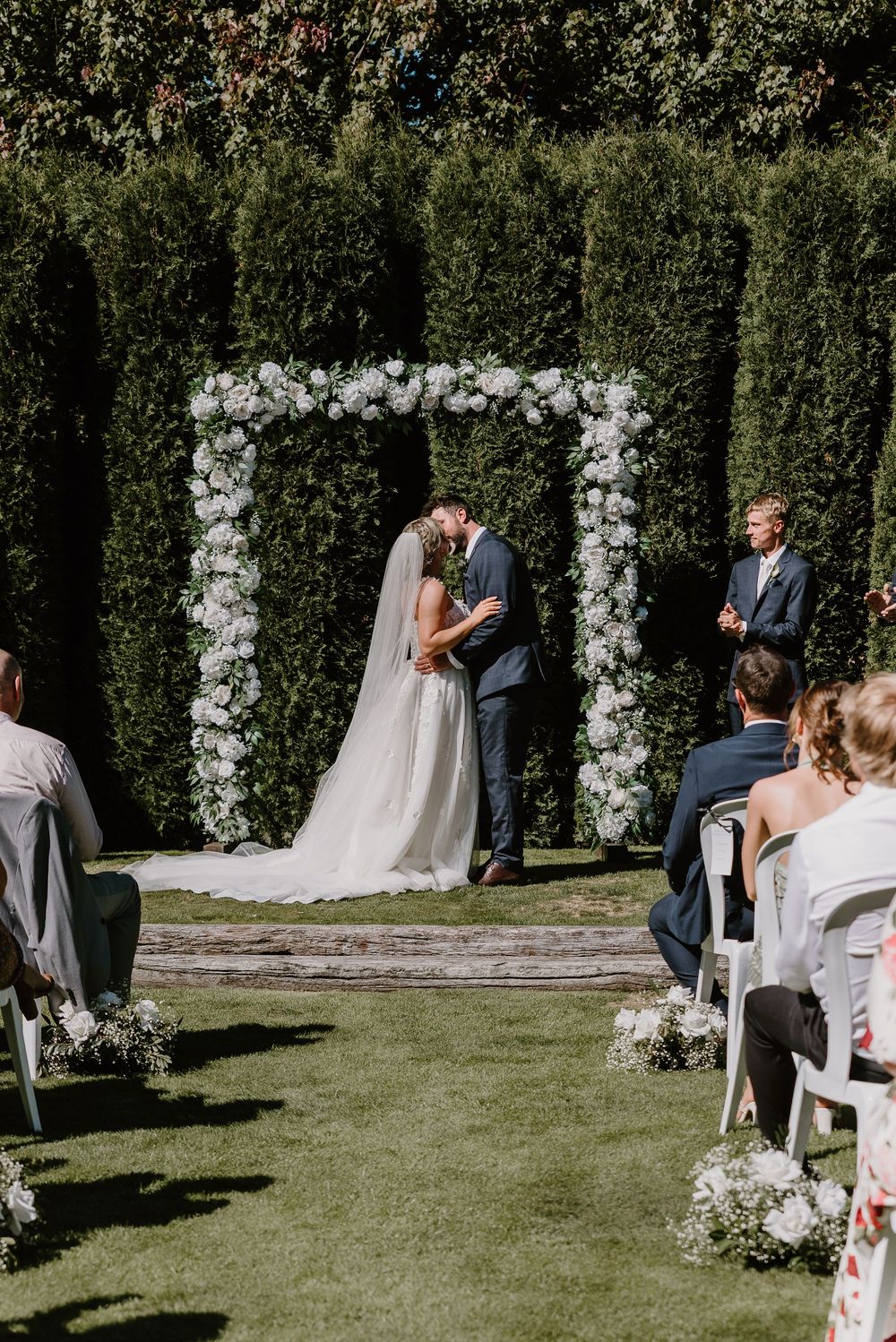 An outdoor wedding ceremony under a white floral arch with guests seated on white chairs.