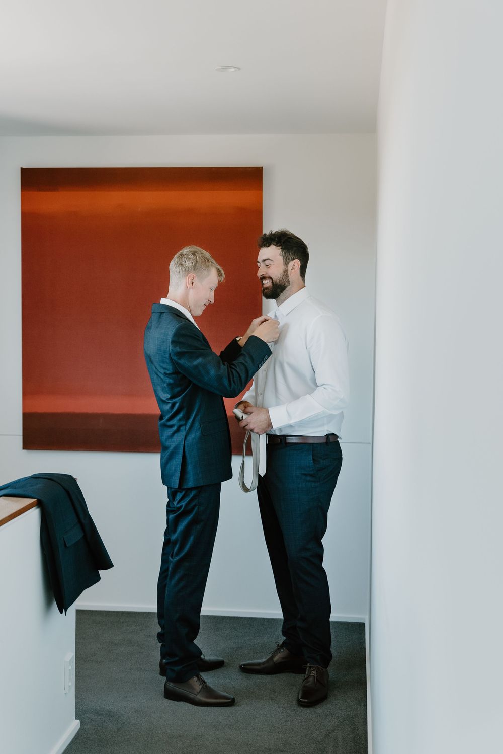 Two men in formal wear helping each other get dressed against an orange wall backdrop.