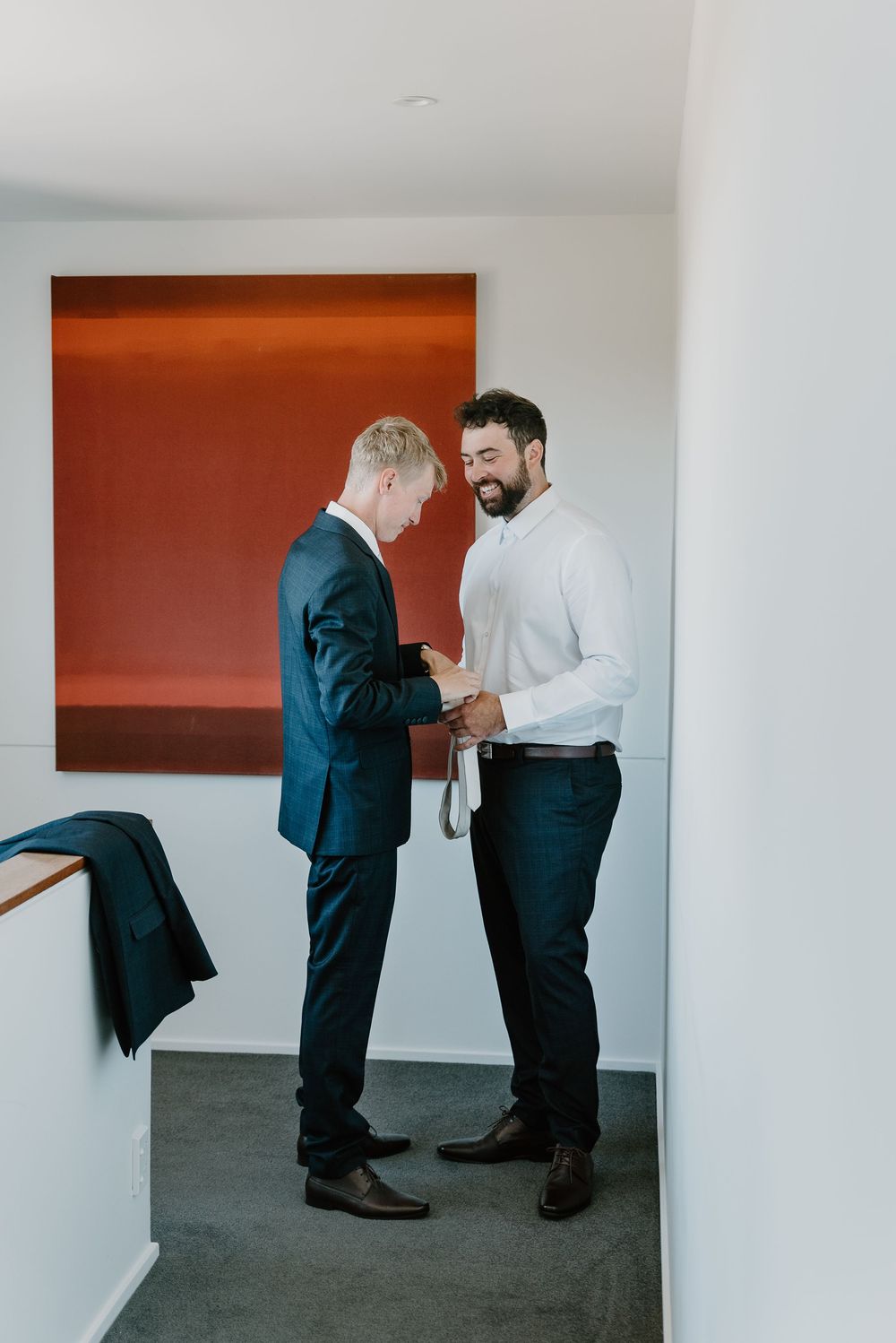 Two men in formal attire adjusting clothing in a room with an orange accent wall and hardwood floors.