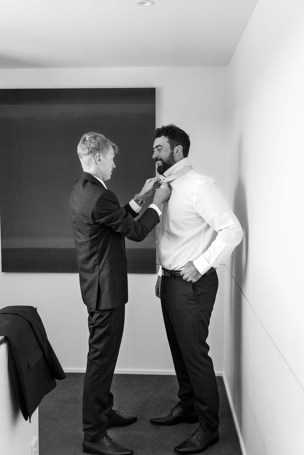 Two dressed-up individuals helping with formal attire adjustments in a minimalist interior setting.