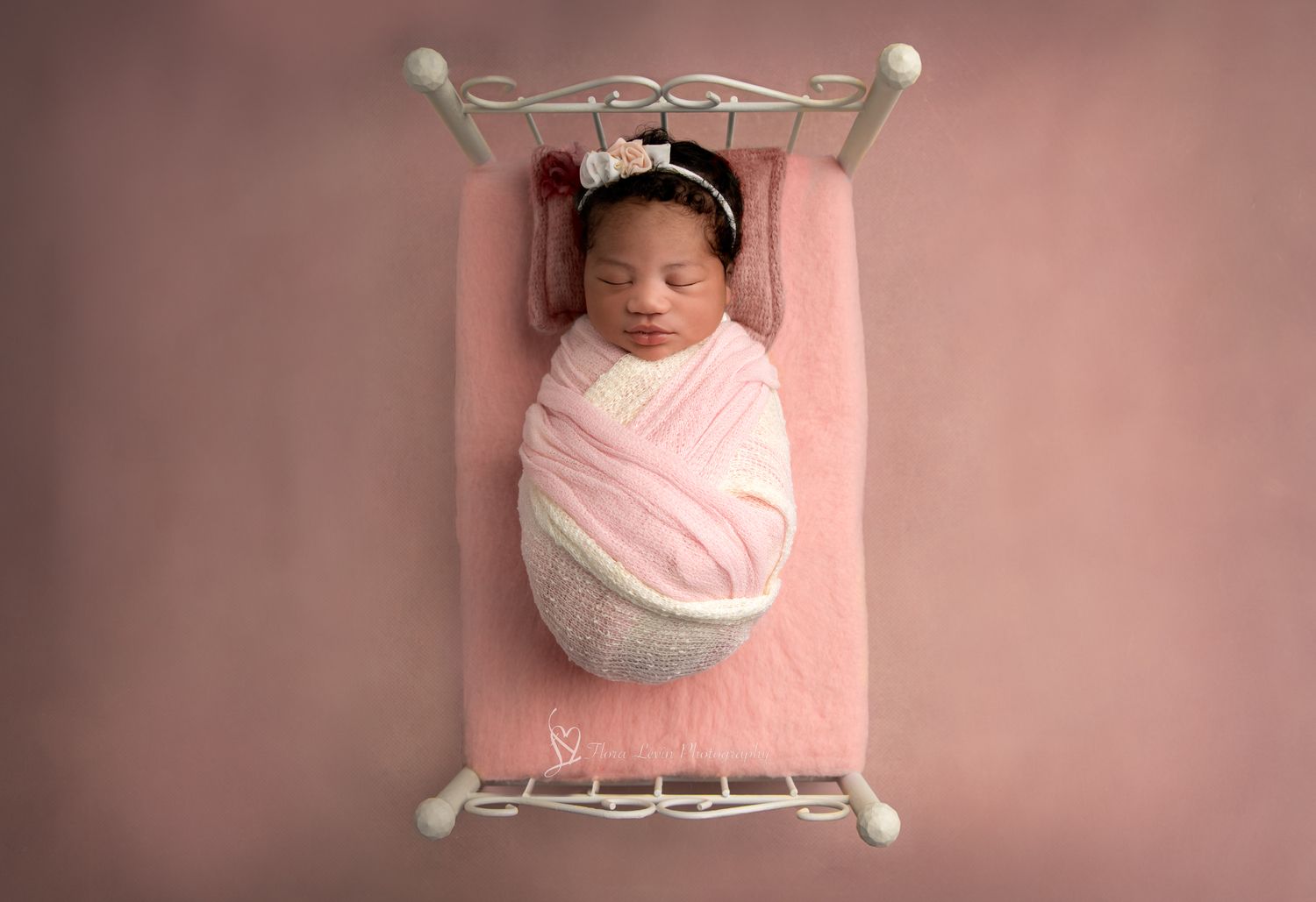 A sleeping newborn baby wrapped in pink and white swaddling cloth lies on a white bed frame with soft pink background lighting.