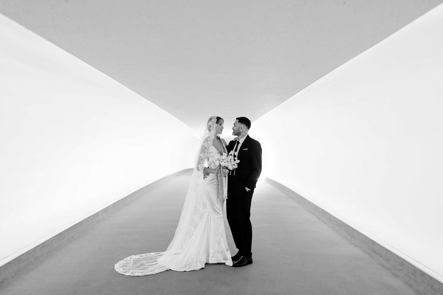 A bride and groom embrace in an elegant pose against a minimalist white architectural corner backdrop.