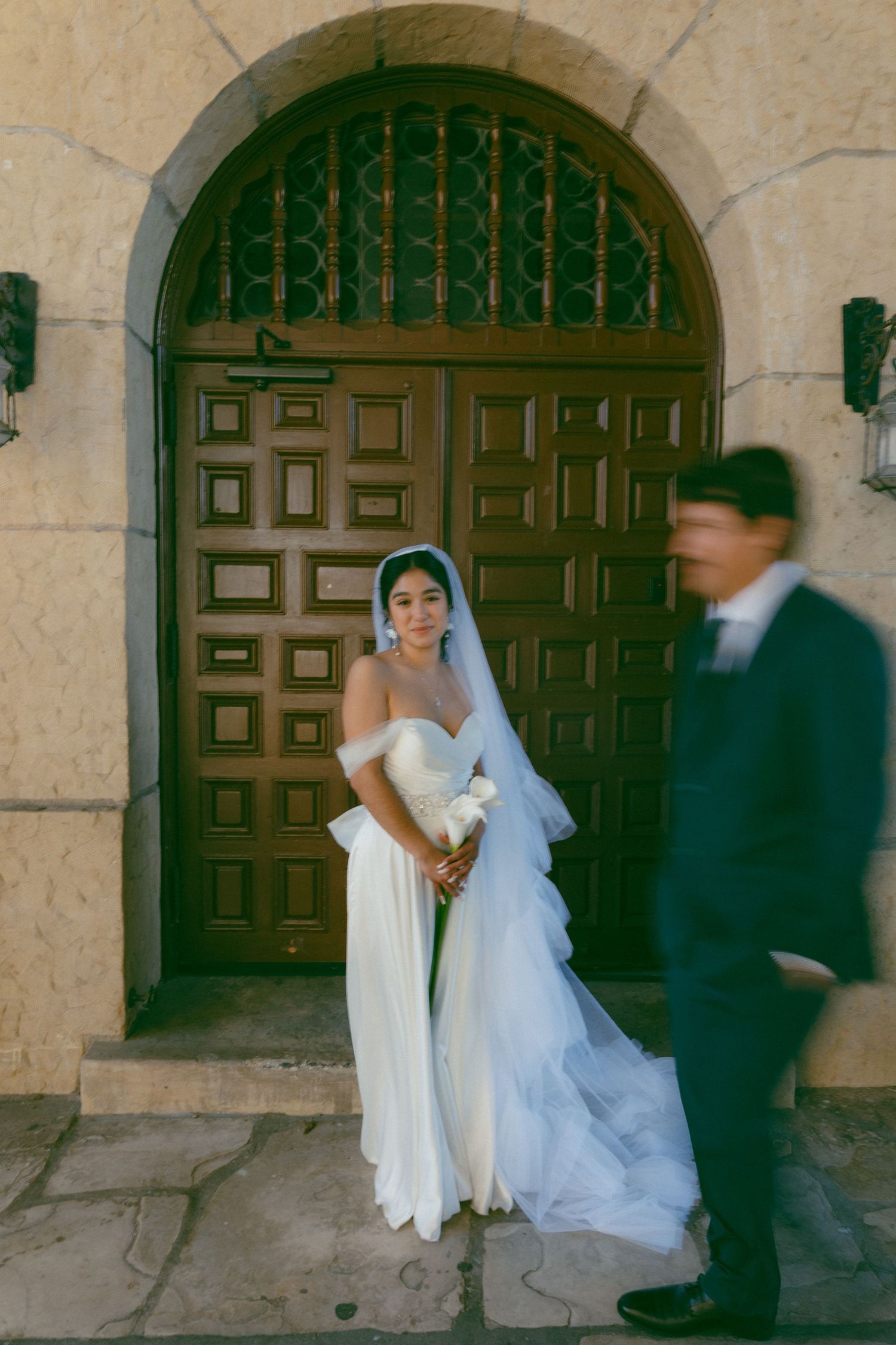 A bride in a white dress and veil poses in front of a grand wooden arched door while a figure walks past.