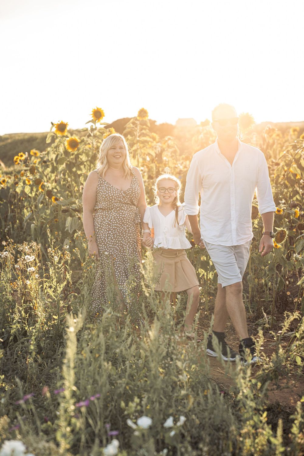 Family walks together through a sunlit wildflower meadow at golden hour during sunset.