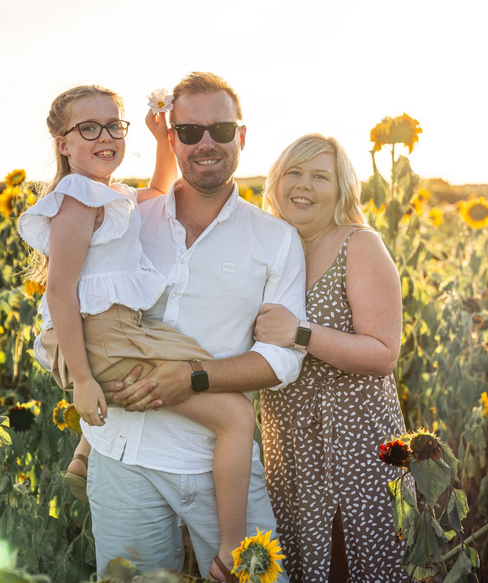 A smiling family poses together in a vibrant sunflower field at sunset wearing casual summer outfits.