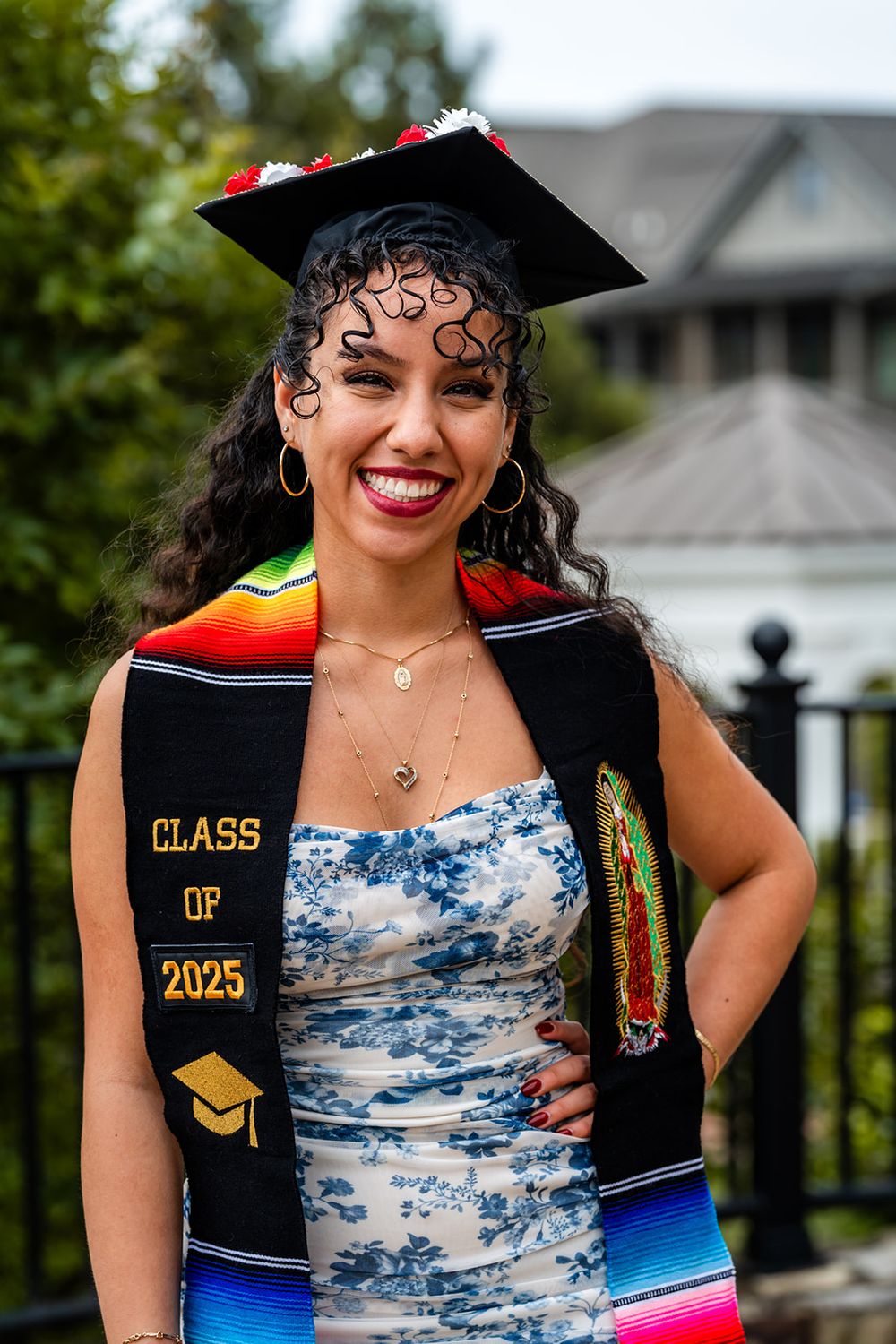 Graduate wearing a black cap and gown with Class of 2025 sash poses in a blue tie-dye dress against outdoor scenery.