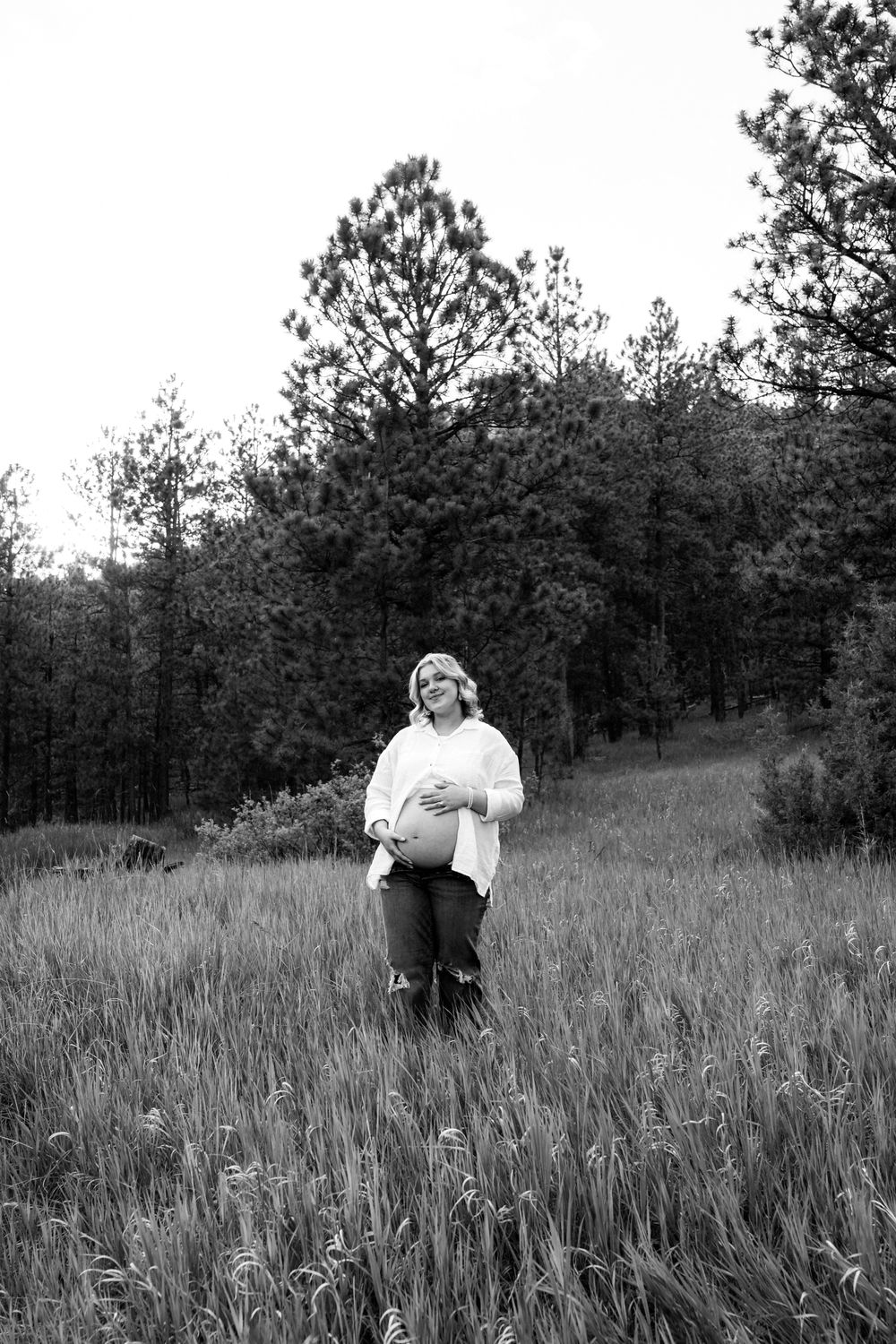 Black and white portrait of person sitting in tall grass field with pine trees in background.