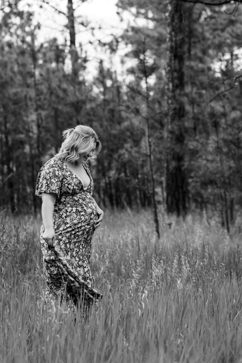 Black and white portrait of someone twirling in a floral dress among tall grass and pine trees.
