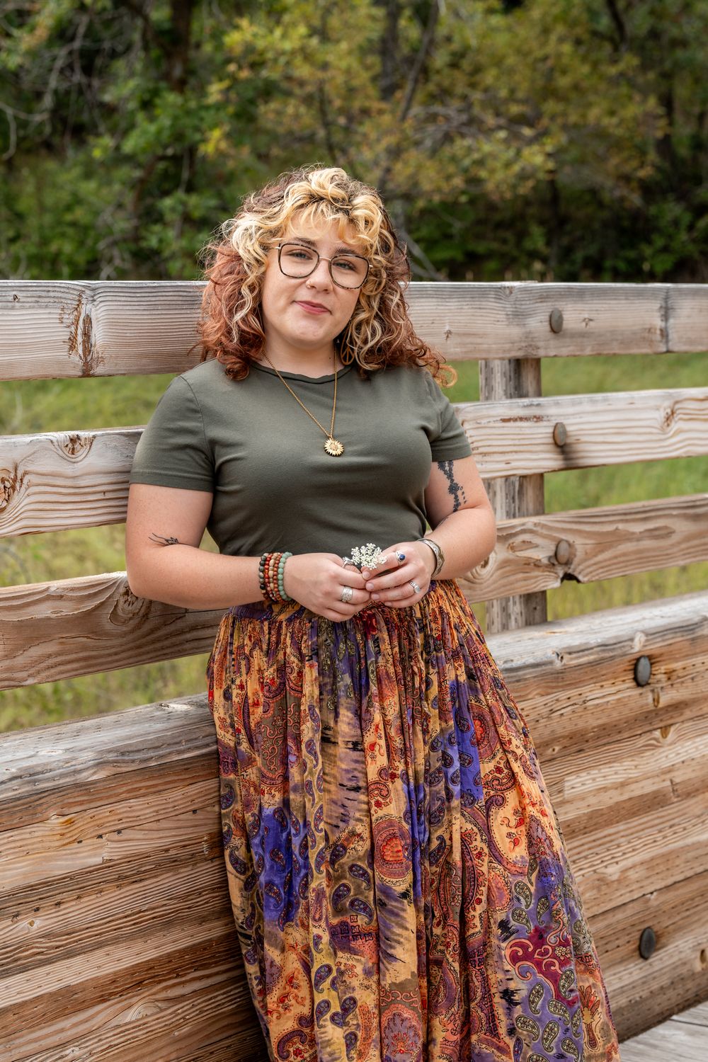 Person sitting on wooden bench wearing floral skirt and olive top outdoors.