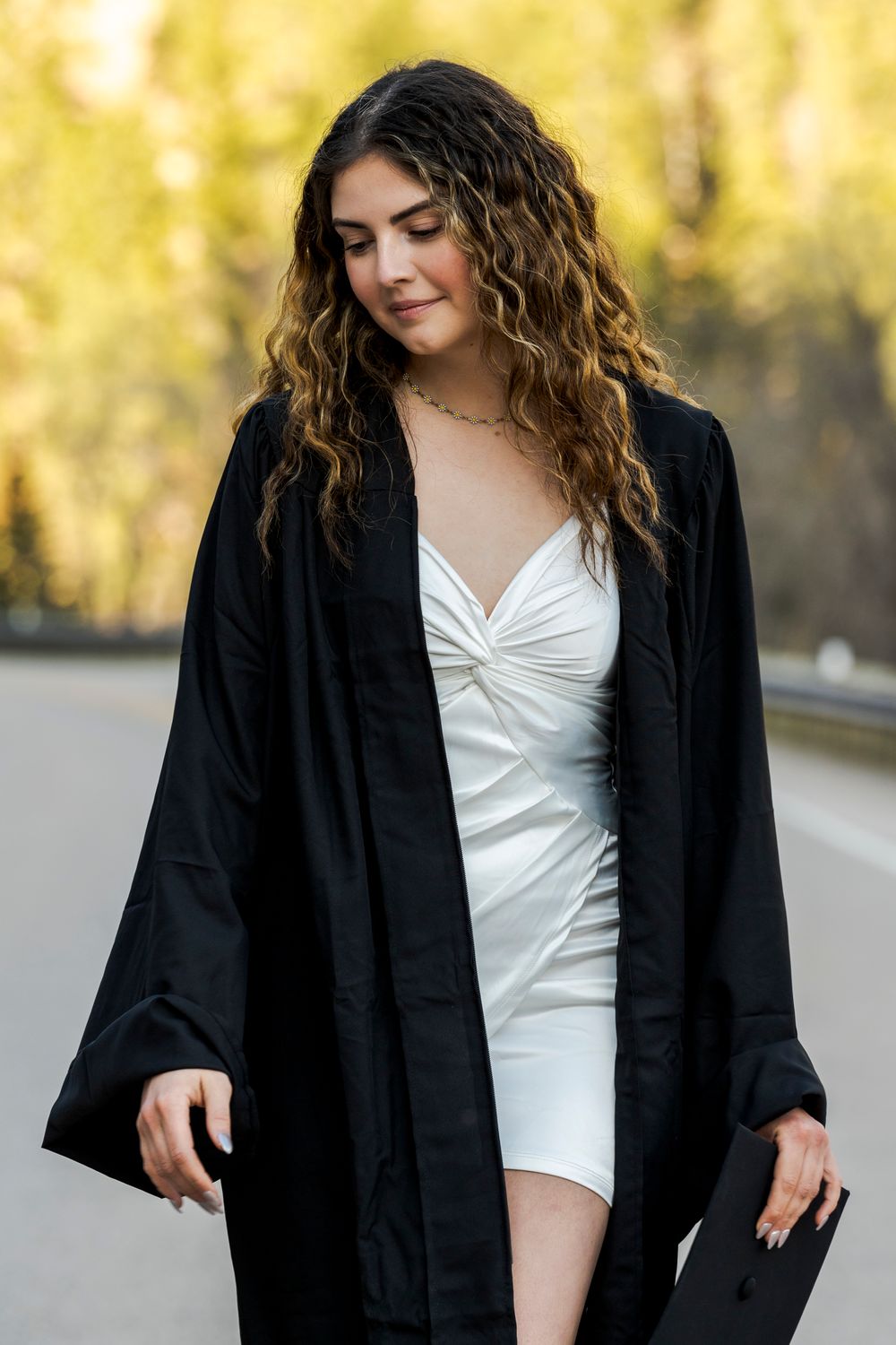 Graduate wearing black graduation gown over white dress in outdoor setting.